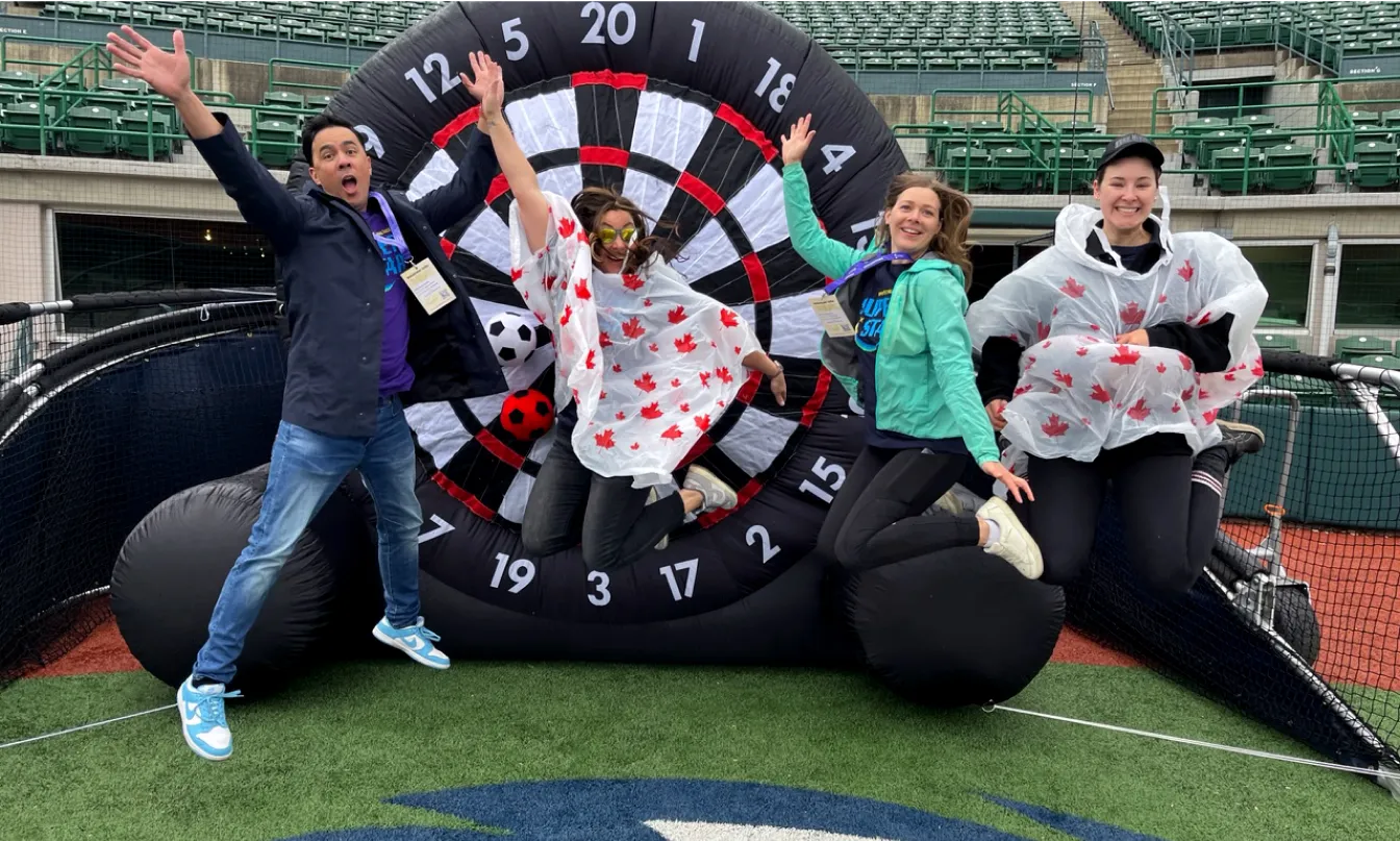 Four people jumping in front of an inflatable dartboard in a sports arena.