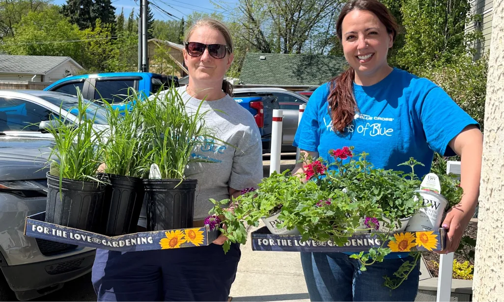 Two women stand in a parking lot, each holding a box filled with potted plants.