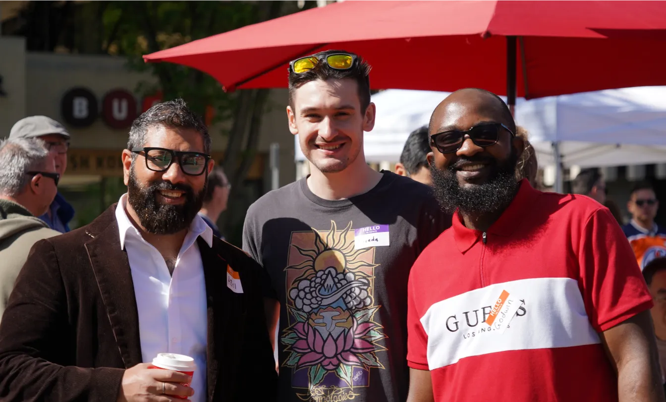Three employees smile while posing for a photo together at an outdoor team event, with a red umbrella in the background.